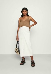 Top cropped castanho com botões, combinado com uma saia midi branca. A modelo usa sandálias pretas e segura uma pequena mala de ombro.