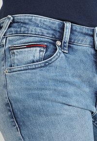 Jeans de mezclilla azul claro con un acabado desgastado, que presentan un pequeño acento a rayas rojas y blancas en el bolsillo frontal y un cierre con botón plateado.