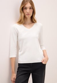 Junge Frau mit welligem blonden Haar, die ein weißes 3/4-Ärmel V-Ausschnitt-Oberteil und schwarze Hosen trägt und an einer hellbeigen Wand steht.