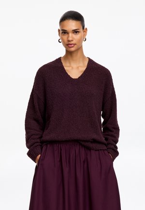 Femme aux cheveux foncés tirés en arrière portant des boucles d'oreilles créoles en or, un pull texturé violet foncé à col en V, et une jupe assortie violet foncé avec les mains dans les poches.