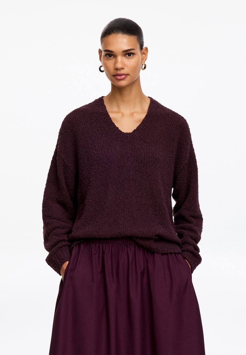 Femme aux cheveux foncés tirés en arrière portant des boucles d'oreilles créoles en or, un pull texturé violet foncé à col en V, et une jupe assortie violet foncé avec les mains dans les poches.