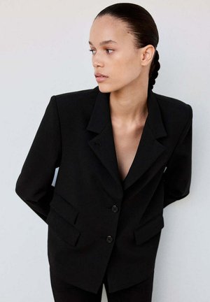 Frau mit geflochtenen Haaren, die einen schwarzen übergroßen Blazer trägt, schaut seitlich vor einem schlichten hellen Hintergrund.