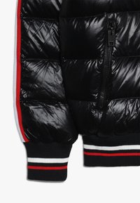 Veste matelassée noire avec une finition brillante, dotée d'une fermeture éclair sur la poche latérale, de poignets côtelés noirs avec des rayures rouges et blanches, et d'un design matelassé.