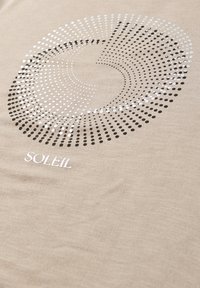 Beżowa tkanina z okrągłym wzorem czarnych i białych kropek tworzących spiralę nad słowem "SOLEIL" napisanym białym tekstem.