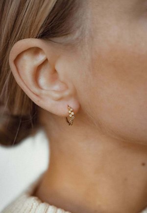 Orecchino a cerchio in oro con un design delicato, caratterizzato da piccole gemme trasparenti disposte attorno alla parte anteriore. Indossato in un orecchio forato, mette in evidenza la sua forma.
