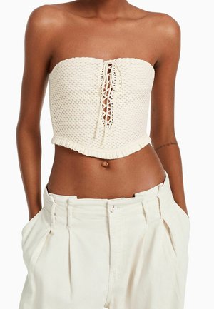 Woman wearing a white textured strapless crop top with lace-up front and high-waisted loose white pants with hands in pockets.