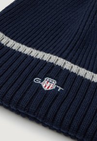 Beanie a coste navy con una striscia grigio chiaro e dettaglio del logo ricamato con accenti rossi e bianchi. Materiale morbido in misto lana.