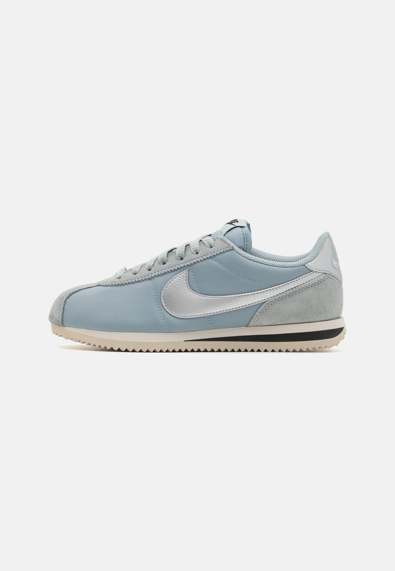 Világoskék Nike sportcipő ezüst swoosh-sal, velúr részletekkel, fehér és fekete talppal, valamint fűzőkkel, oldalnézetből, sima háttér előtt.