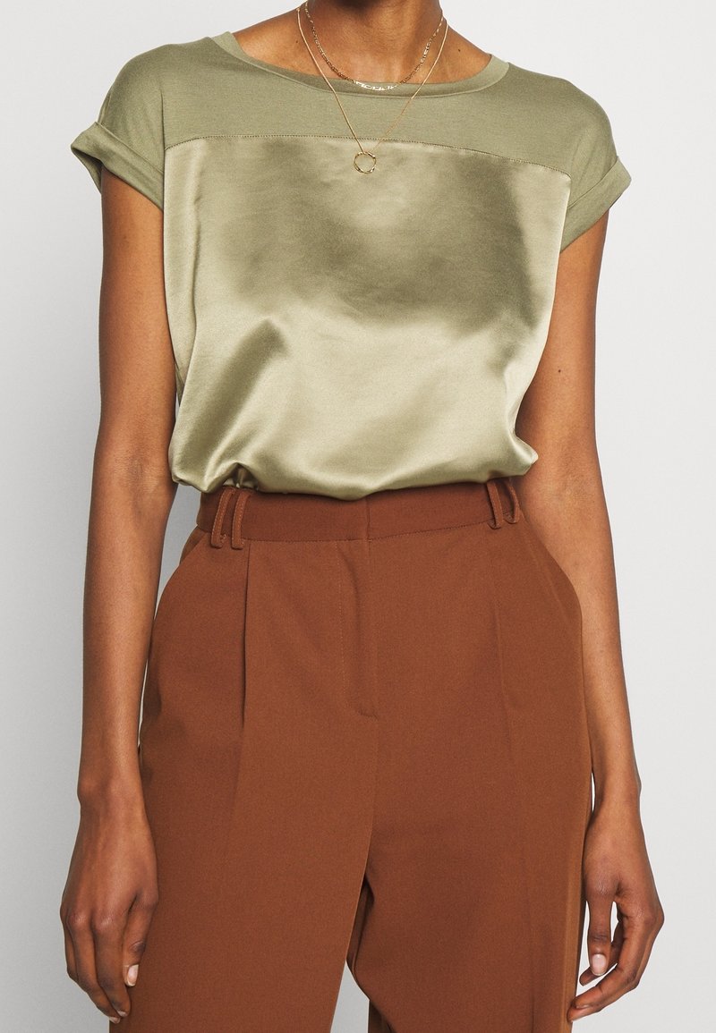 Haut vert olive à finition satinée, avec un col arrondi et des manches retroussées, associé à un pantalon marron taille haute.