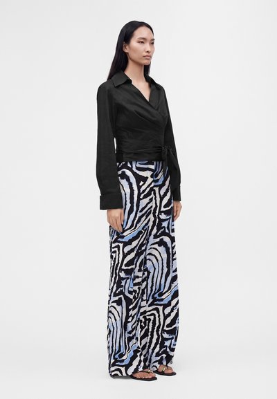 Femme debout portant un chemisier cache-cœur noir et un pantalon à jambes larges avec un imprimé zèbre abstrait noir, blanc et bleu et des sandales noires.