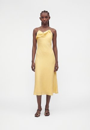 Nainen seisoo päällä keltainen hihaton midimekko, jossa on laskostettu kaula-aukko ja ohuet olkaimet, yhdistettynä mustiin avoimilla varpailla sandaaleihin.