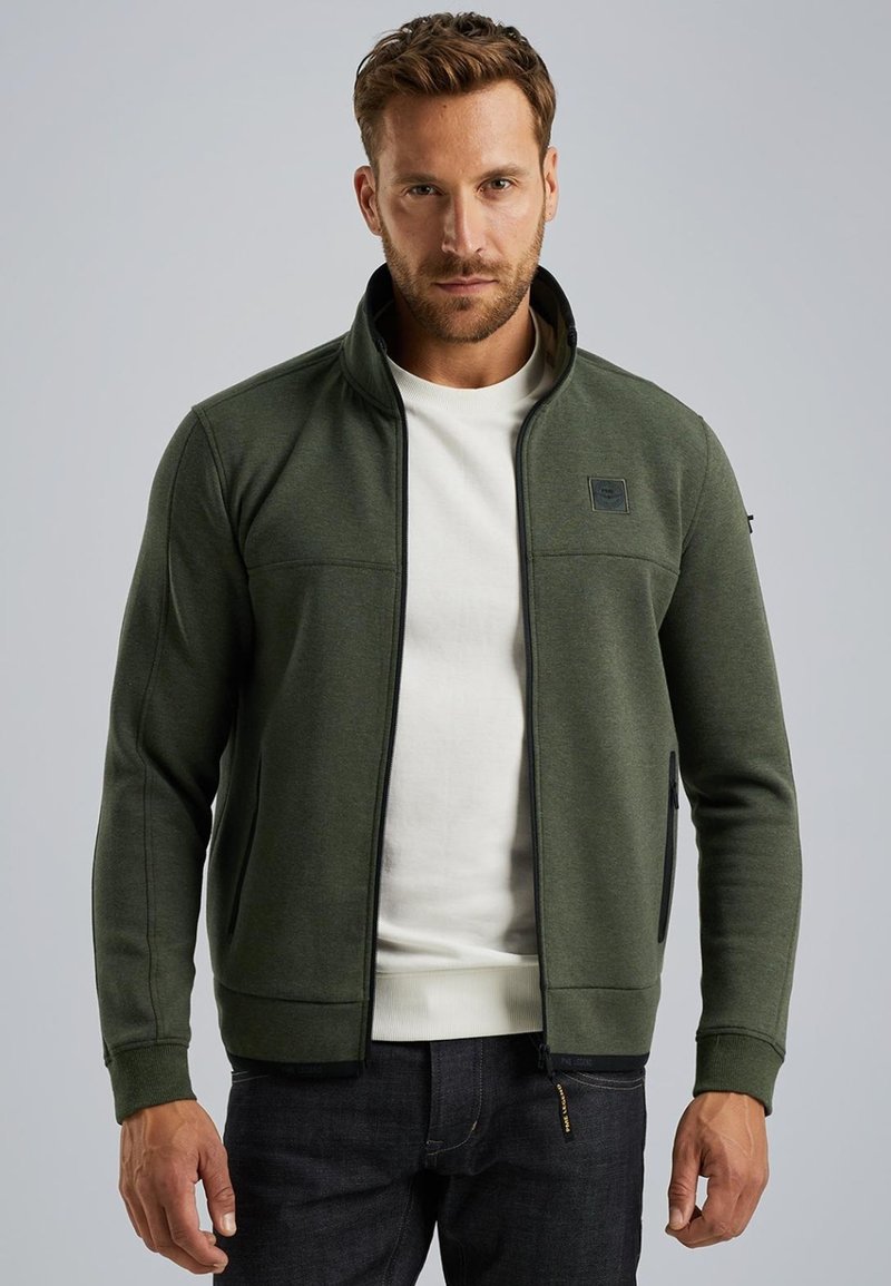 Giacca verde con zip realizzata in tessuto morbido, dotata di un colletto alto, pannelli testi e tasche laterali. Indossata sopra una camicia bianca.