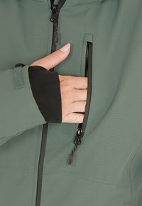 Grågrön softshell-jacka med en dragkedjeficka, med svarta elastiska ärmslut och en slät yta. Hand som är placerad inuti fickan.