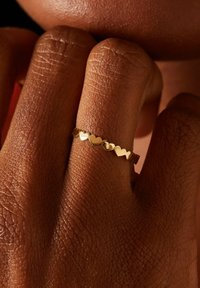 Gouden ring met een golvend ontwerp en hartvormige uitsnijdingen. De band is glad en reflecterend, in contrast met een textuur van de huid.