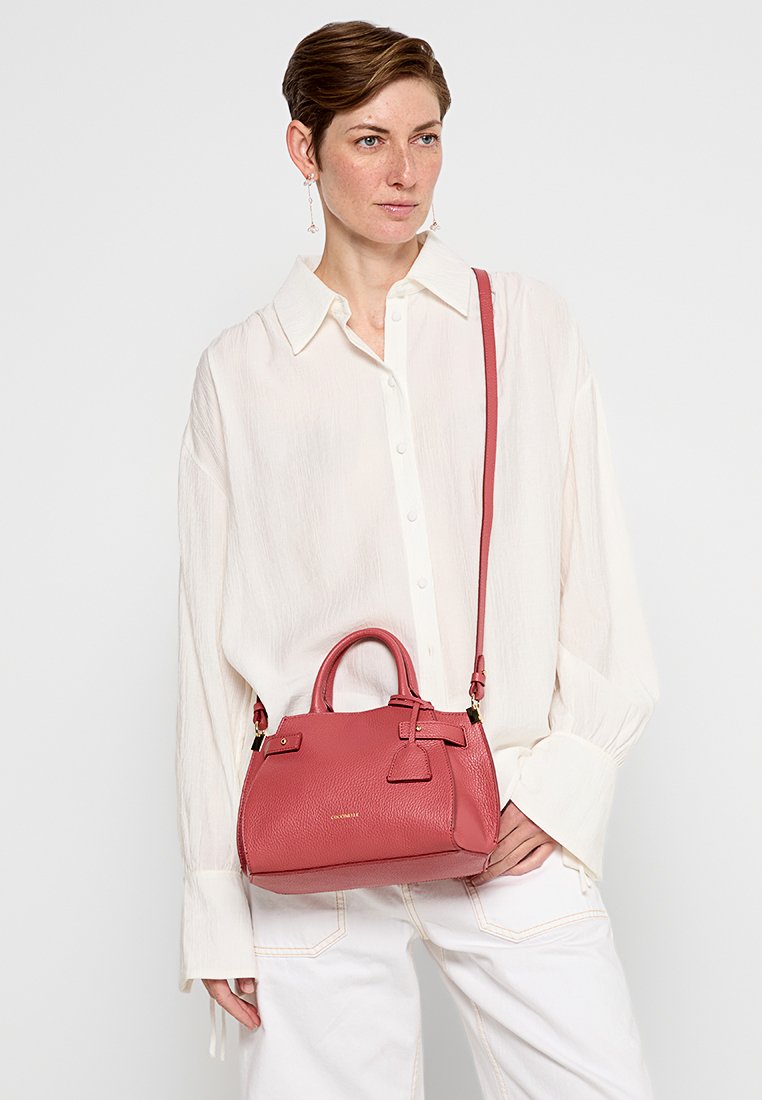 Piccola borsa a mano in pelle rosa con una forma strutturata, doppi manici superiori e una tracolla rimovibile. Caratterizzata da una superficie testurizzata e un sottile logo dorato.