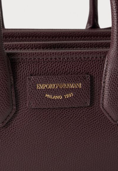 Gros plan d'un sac en cuir marron foncé texturé montrant une plaque logo dorée Emporio Armani Milano 1981 et une partie des deux poignées.