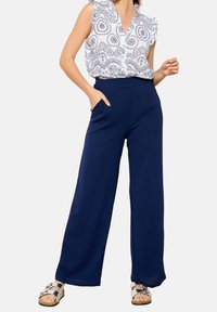 Námořnické široké kalhoty s hladkou texturou, vybavené bočními kapsami a vysokým pasem. V kombinaci s bílou vzorovanou blůzou a stříbrnými sandály.