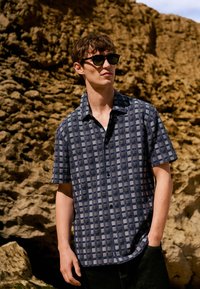 Uomo con occhiali da sole neri e camicia a maniche corte blu a quadri, in piedi davanti a uno sfondo roccioso all'aperto.