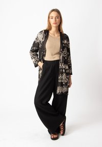 Ilgas juodas kimono su sudėtingais sidabro ir aukso raštais, dėvimas virš smėlio spalvos banguoto audinio marškinėlių ir juodų plačių kelnių, turinčių laisvą siluetą.