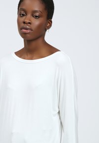 Maglietta bianca a maniche lunghe con un ampio scollo a barca, texture morbida e vestibilità rilassata. Il tessuto liscio cade in modo fluido, evidenziando il comfort.