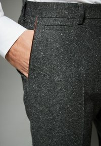 Pantalones de lana texturizada de color gris oscuro, con un corte ajustado, que presentan un bolsillo lateral y detalles de costura mínimos. La tela tiene un patrón moteado sutil.