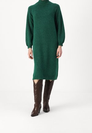 Femme portant une longue robe pull col roulé verte et des bottes en cuir marron montant jusqu'aux genoux, debout sur un fond blanc uni.