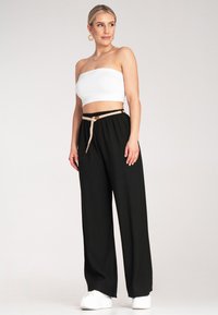 Czarne spodnie o szerokich nogawkach z elastycznym pasem na sznurek. Połączone z białą bluzką bez ramiączek. Modelka stoi w sneakersach, na tle neutralnym.