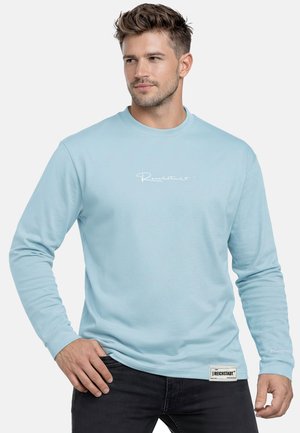 Mann trägt ein hellblaues Langarmshirt mit weißem Text auf der Brust und einem rechteckigen Etikett nahe der Taille, steht mit einer Hand auf der Hüfte.