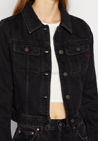 Kvinna som bär en figursydd svart denimjacka med bröstfickor och knappar över en vit kortärmad topp, kombinerad med svarta denimjeans.