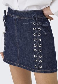 Jupe en denim mini avec des détails à lacets sur les côtés, présentant des œillets en argent et des coutures sombres. Texture lisse et couleur bleue classique.