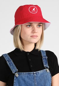 Cappello a secchiello Kangol rosso con una visiera larga, caratterizzato da un logo circolare. Tessuto liscio con una texture interna bianca contrastante.