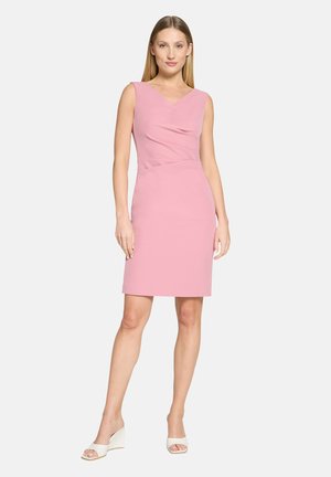 Kobieta stojąca, ubrana w różową sukienkę bez rękawów sięgającą kolan oraz białe sandały na koturnie z odkrytymi palcami, z prostymi blond włosami na jednolitym jasnym tle.