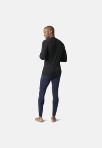 Sort langærmet skjorte og marineblå leggings, tætsiddende Design, glat tekstur, båret af en mand, der står barfodet, set bagfra.