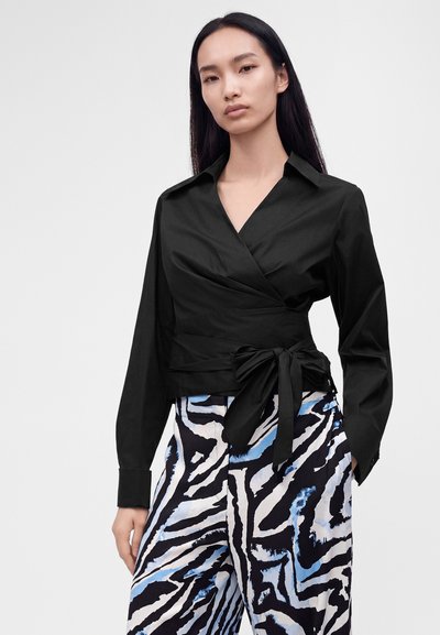 Femme aux longs cheveux noirs portant un chemisier noir croisé noué à la taille et un pantalon taille haute à motif zèbre bleu et noir.