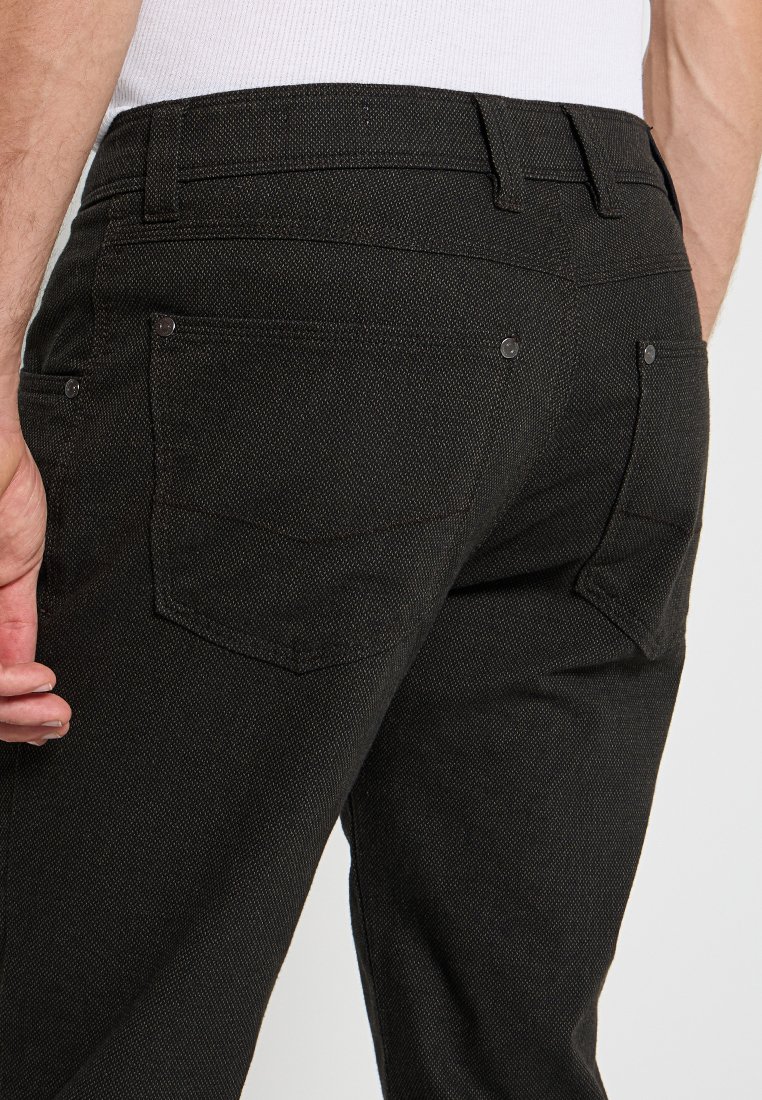 Mann in schwarzen strukturierten Hosen und weißem Hemd, von der Taille abwärts gezeigt, mit der rechten Hand entspannt an seiner Seite.