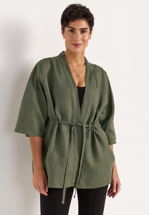 Woman with short dark hair wearing an olive green belted linen jacket over a black top and black pants, standing against a white background.