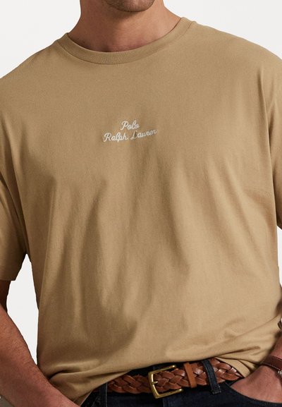 Vyras, dėvintis šviesiai rudos spalvos Polo Ralph Lauren marškinėlius, įstatytus į tamsias džinsus, su ruduoju pintu odiniu diržu ir auksiniu sagtimi.