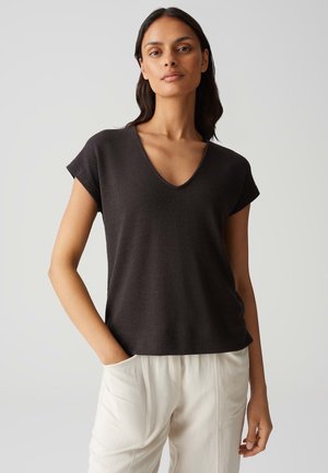 Top grigio scuro a maniche corte con scollo a V, realizzato in un tessuto morbido e testurizzato. Il modello lo indossa con pantaloni color crema, con le mani nelle tasche.