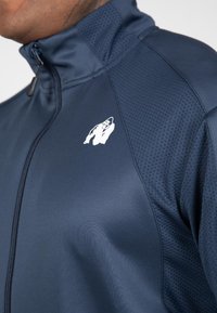 Marine blauwe zip-up jas met een gestructureerd patroon, gladde stof en een wit gorilla-logo op de bovenlinkerkant.