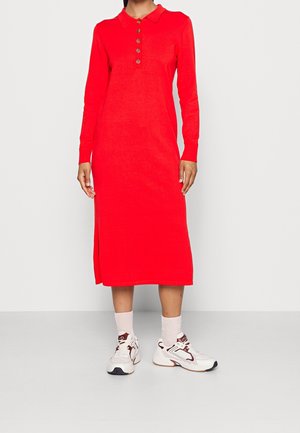 Kvinde iført en rød midi-kjole med lange ærmer, knapper og krave, kombineret med hvide og burgunderfarvede sneakers samt hvide sokker.