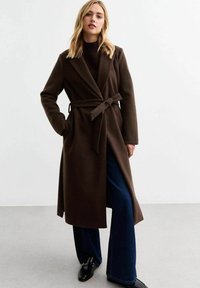 Bruine, lange wikkeljas met een riem, voorzien van eengetailleerde kraag en zijzakken, gecombineerd met donkere, uitlopende spijkerbroek en glanzende zwarte schoenen.