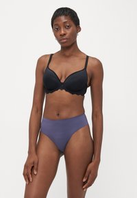 Zwarte bh met kantafwerking en gevoerde cups. Gecombineerd met hoog getailleerde, gladde paarse slipjes. Neutrale achtergrond.