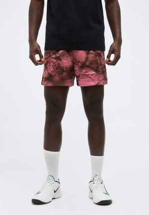 Bărbat purtând pantaloni scurți sport cu model roz și negru, șosete albe până la gleznă, adidași albi Nike și un tricou negru, stând pe un fundal alb.