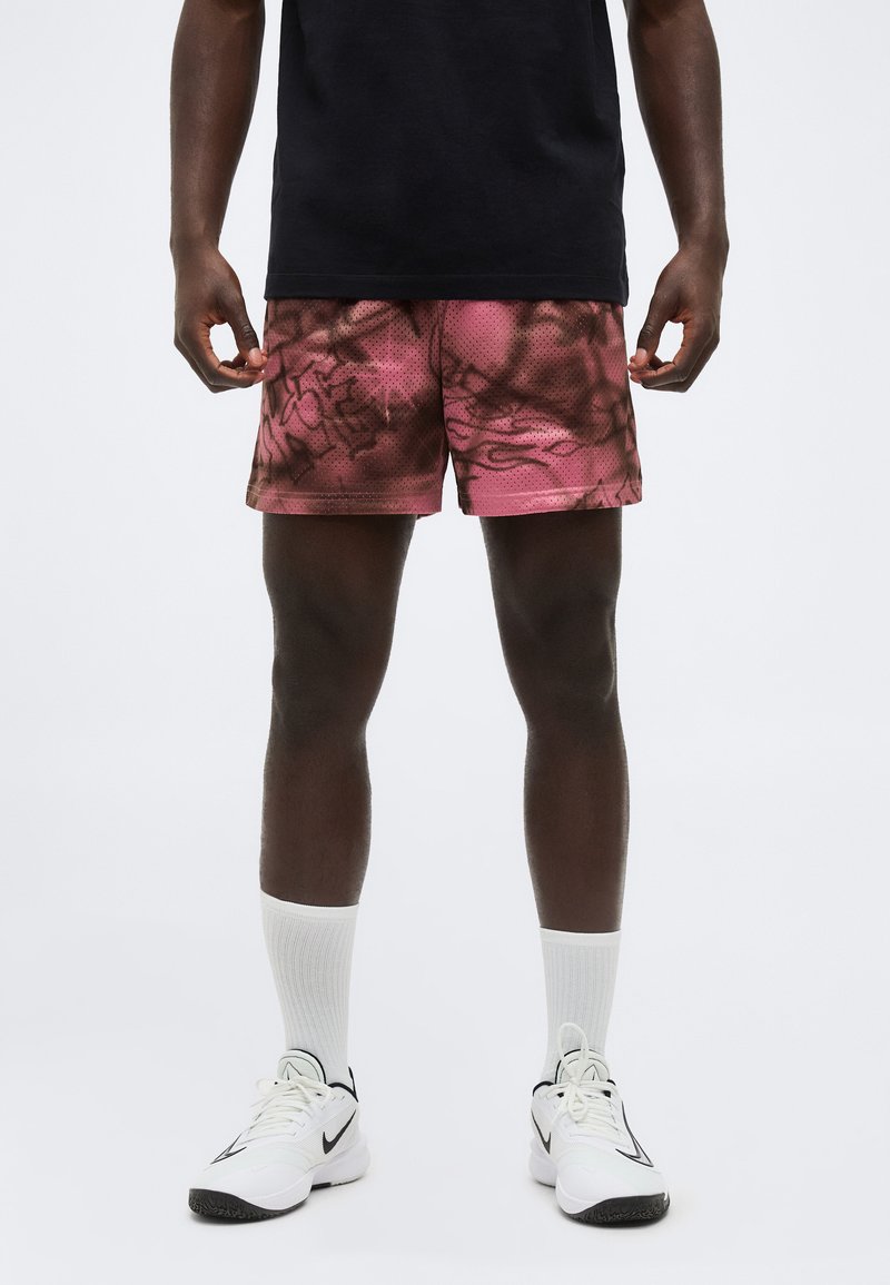 Άνδρας που φοράει ροζ και μαύρα αθλητικά σορτς με σχέδια, λευκές κάλτσες πλήρους μήκους, λευκά αθλητικά παπούτσια Nike και μαύρο πουκάμισο, στέκεται μπροστά σε λευκό φόντο.