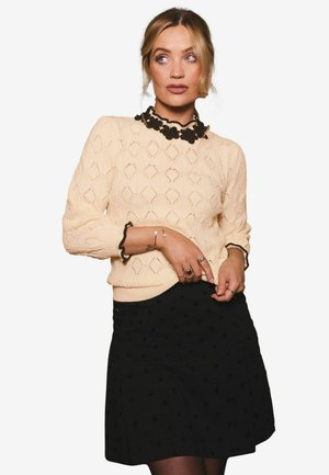 Maglione lavorato a maglia di un beige chiaro con motivi a rombo, colletto florale nero e gonna nera con pois; aderente con maniche lunghe e bordi arricciati.