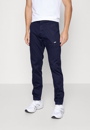 Mężczyzna ubrany w granatowe spodnie cargo, białe trampki i białą koszulkę, stojący na neutralnym tle.