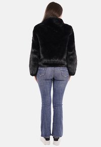 Giacca nera in pelliccia sintetica con colletto alto, progettata con maniche lunghe, abbinata a jeans svasati blu e sneakers bianche.
