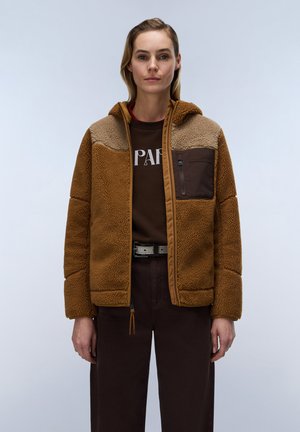Donna che indossa una giacca in pile marrone e beige sopra pantaloni e camicia marrone scuro con lettere stampate, in piedi contro uno sfondo chiaro e uniforme.