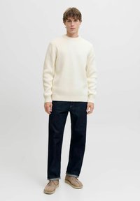 Maglione a costine crema con collo a giroclassico, abbinato a jeans in denim scuro e scarpe in suede beige. Presenta maniche lunghe e una vestibilità rilassata.