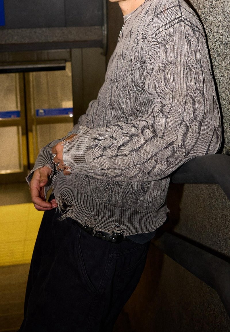 Personne appuyée contre un mur texturé portant un pull gris en maille torsadée avec des poignets usés et un pantalon sombre.